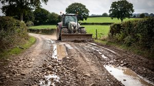Chemin rural empierré dégradé avec ornières et flaques, bordé de haies bocagères, tandis qu’un tracteur agricole équipé d’une lame niveleuse prépare sa remise en état.