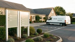 Entrée soignée d’une entreprise rurale avec panneaux directionnels sans inscription, parking visiteurs aménagé et véhicule de livraison, éclairée par une lumière naturelle.
